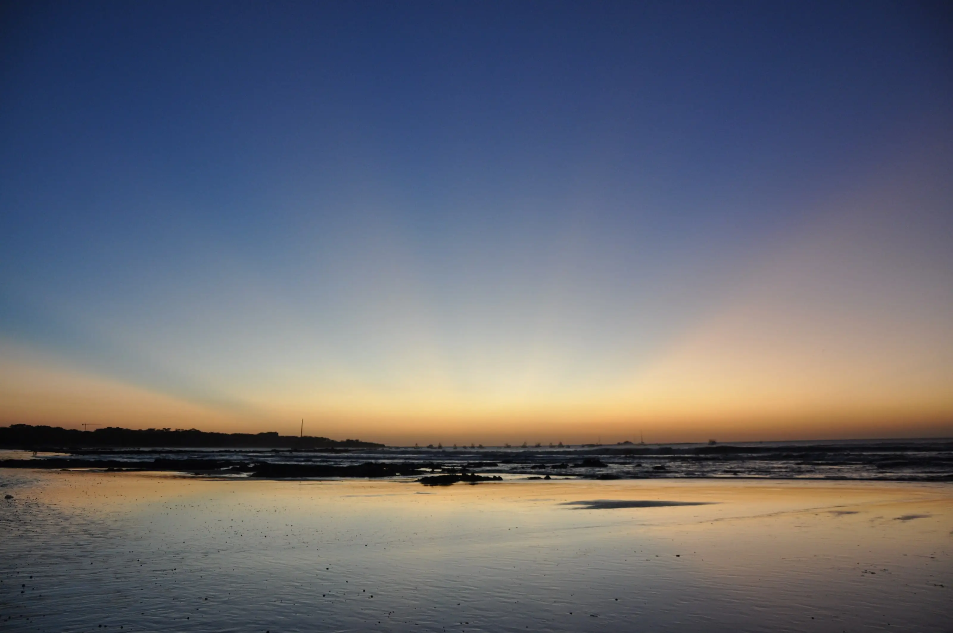 Twilight over Playa Tamarindo: a clear sky fading from deep blue to orange at the horizon, boats anchored in the bay and wet sand reflecting the glow at low tide.